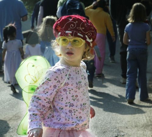 Young girl in a fairy costume with pink floral top, yellow glasses, and green wings walking in a sunny outdoor crowd.